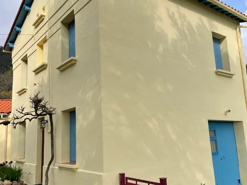 Façade de maison en enduit jaune pâle aux volets bleus, après ravalement