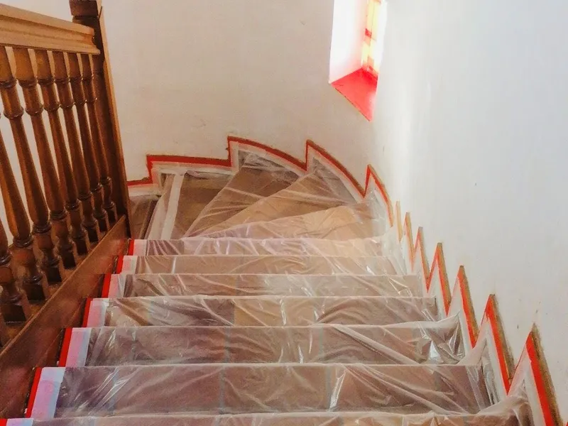 Escalier vu d’en haut, marches couvertes d’un film plastique bordé de ruban de masquage, rampe en bois et murs blancs