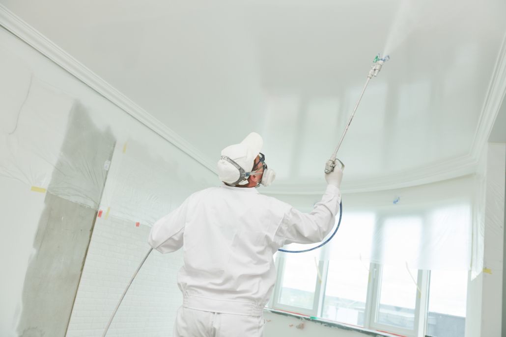 Peintre en combinaison et masque appliquant la peinture au plafond au pistolet