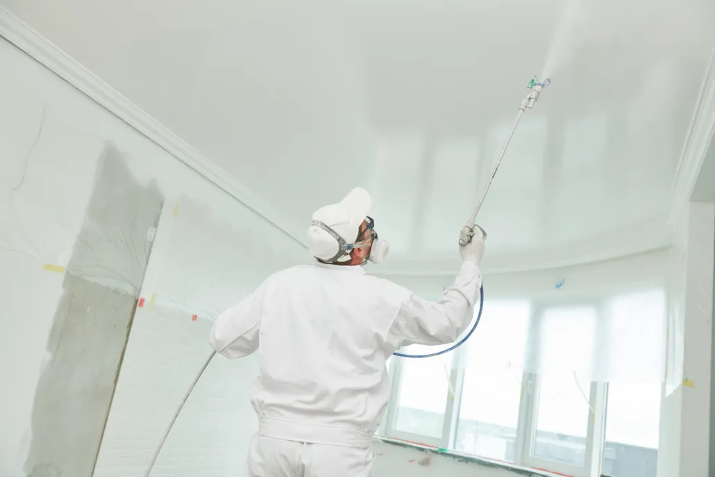 Peintre en combinaison et masque appliquant la peinture au plafond au pistolet